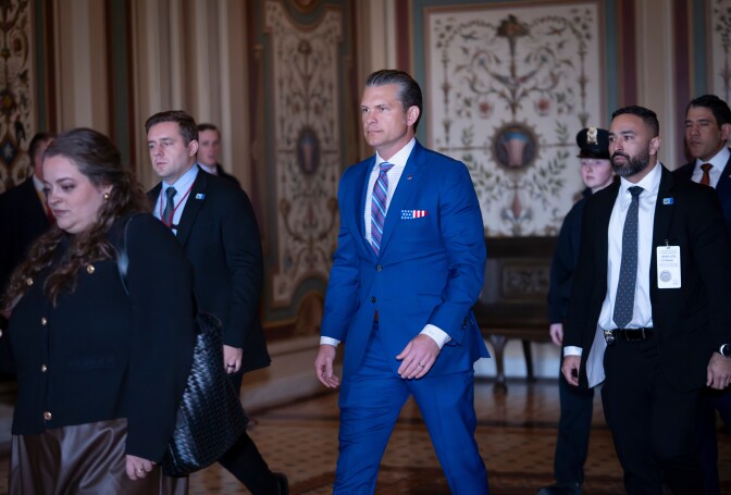 A white man in a slim-cut blue suits walks in a corridor near other people.