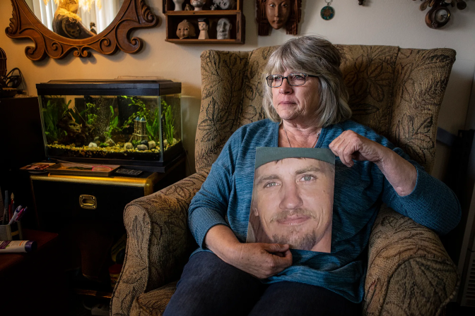 A woman with graying shoulder length care and eyeglasses is seated in a wingback chair holding a large photo of a young man with light skin, green eyes, a mustache and goatee. A fish tank is beside her on a table.
