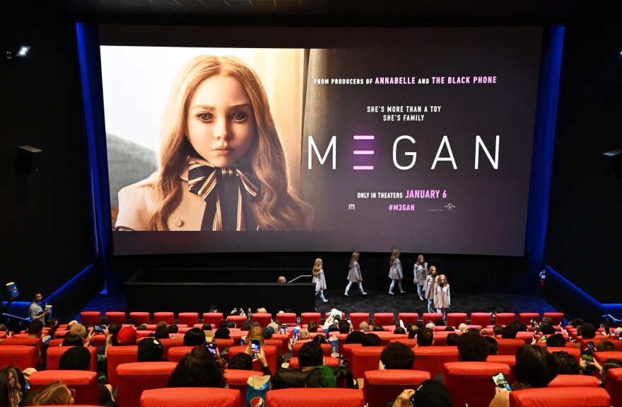 People sit in a movie theater facing a large screen that shows a young robot girl with the title "MEGAN."