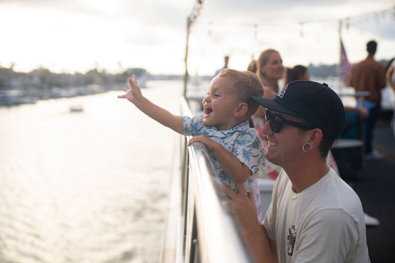 A father holds his young son on a boat, high enough to see over the railing. The son is waving to someone outside of the frame. Both he and his dad are smiling.
