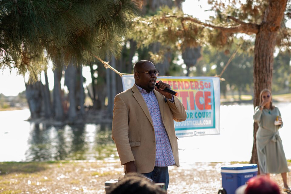  Superior Court judge candidate George Turner speaks at a Defenders of Justice campaign event. He wears a tan suit and speaks into a wireless microphone. There are trees and a body of water behind him. 