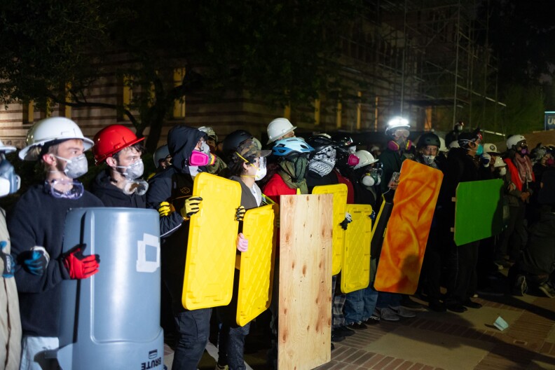 A row of people wearing helmets and masks hold makeshift shields some made out of cut up trash cans, others bin lids, and wooden boards.