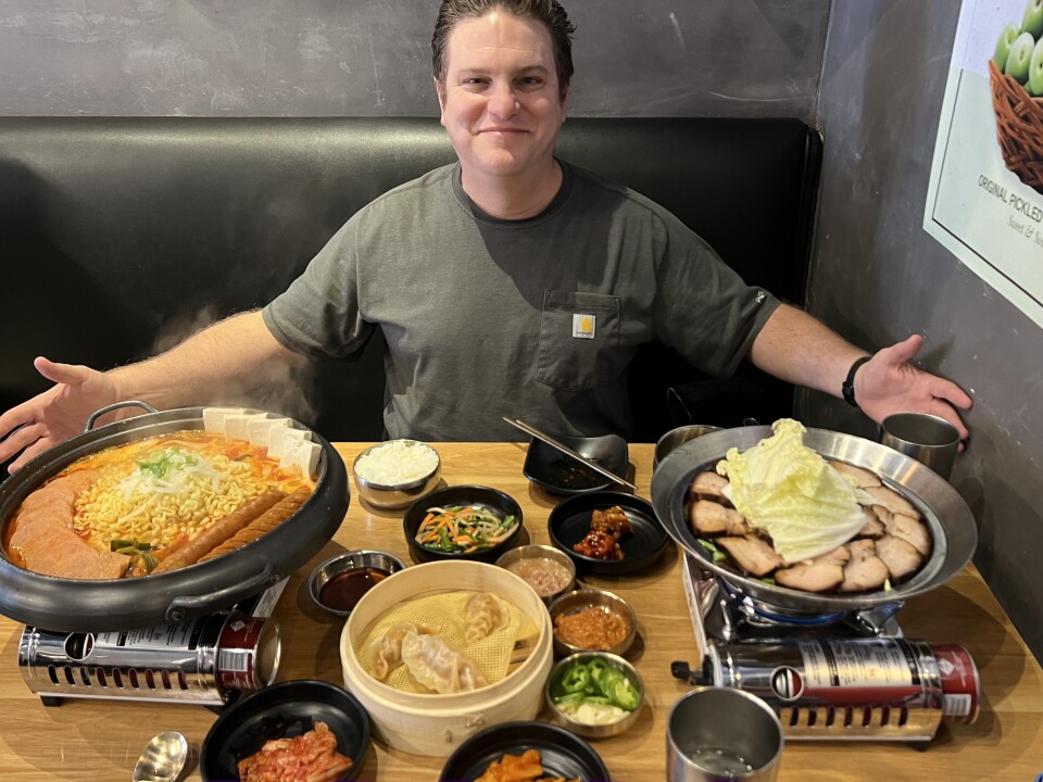 A man with a light skin tone, wearing a grey t-shirt is sitting in a black vinyl booth with arms outstretched and smiling. In front of him are two large portable burners. One carries a large bowl of soup, while the other has slices of meat. In between the burners, there are various small dishes filled with different types of banchan.