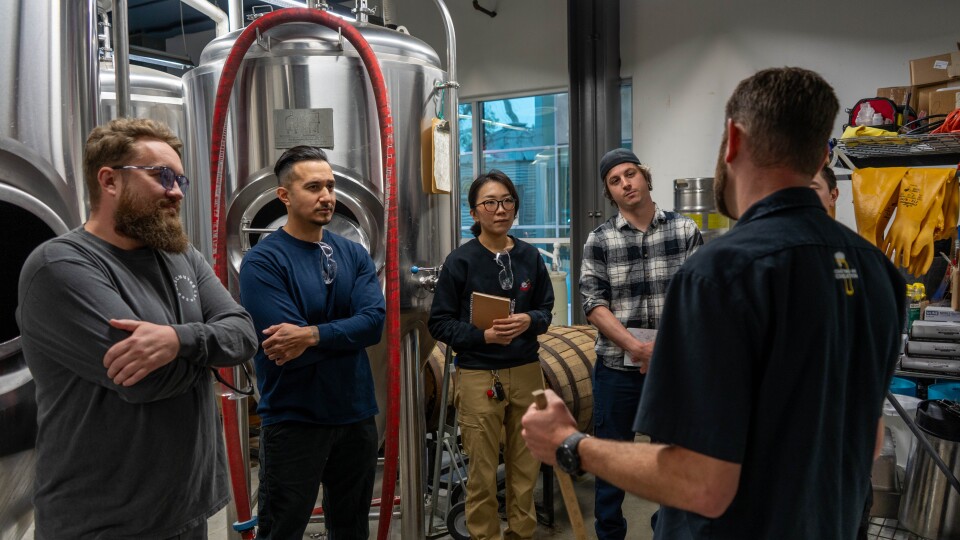 Five people stand next to metal vessels larger than them. Four look at one, who is talking to them.