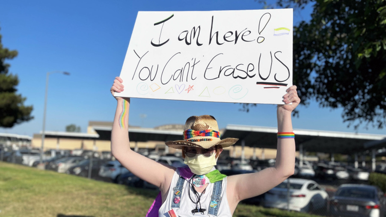 A light-skinned person wearing a yellow surgical mask and a straw hat with a rainbow-colored band holds up a sign that reads "I am here! You Can't Erase Us"