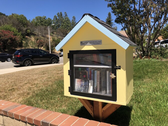 A small yellow box with a clear glass door that resembles a house with blue trim on the base of the pointed roof sits outside in front of a short wall topped with red brick. The box contains two shelves full of books. On the second shelf, one book sits on its side with the cover facing out lying on its side. Behind the library is a blue sky above a small hillside with greenery. In the foreground are two black SUV cars parked along the street. 