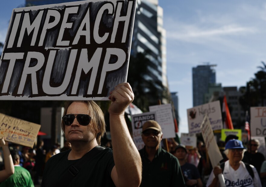 A man with light-tone skin and sunglasses holds a handmade sign that reads: Impeach Trump. Other sign-carrying protesters are in the background.