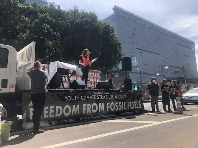 A young woman stands on a stage with a banner "Freedom From Fossil Fuels"