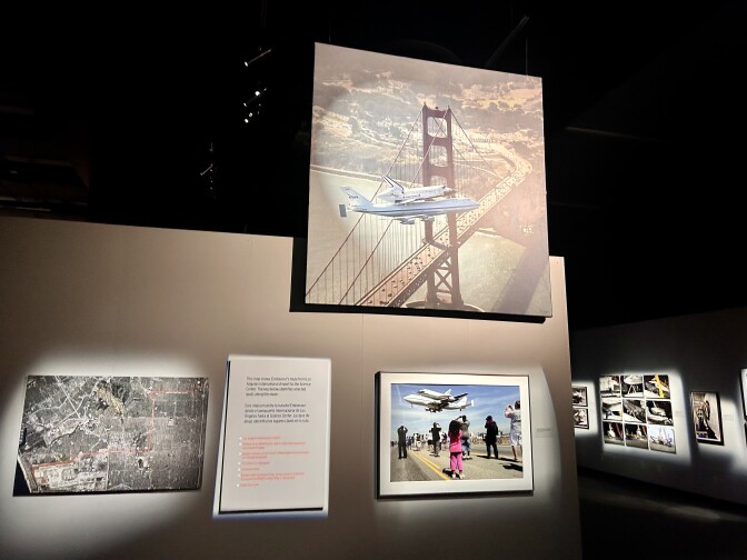 The interior of a photo gallery in a museum. The largest photo towards the top of the wall shows a space shuttle being flown near the Golden Gate Bridge in San Francisco, and the smaller photo just below it shows several people, including a child wearing all pink, watching the space shuttle from a street.