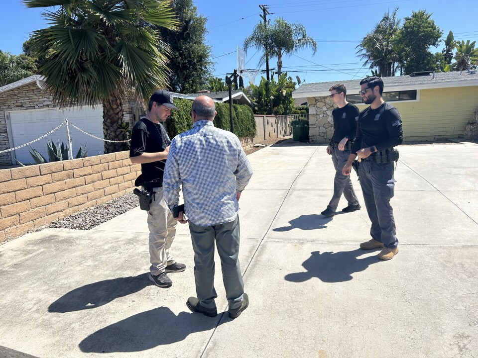Three men wearing black shirts with gun holders stand around a driveway next to a man with a button up shirt and slacks who's back faces the camera. 