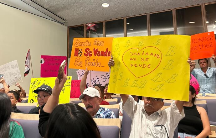 A group of residents it in blue chairs in Santa Ana City Hall holding brightly colored signs that read "Santa Ana no se vende" or "Santa Ana is not for sale," and "Santa Ana no se vende, se cuida" or "Santa Ana is not to sell, its to take care of." 