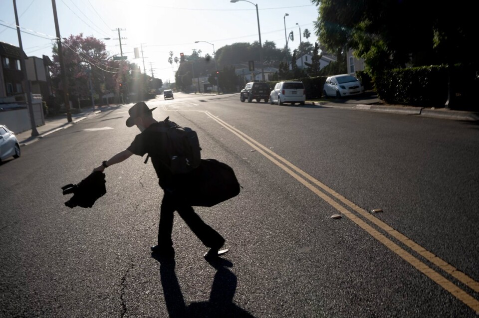 A man in a hat, carrying a bag, walks across the street in silhouette.