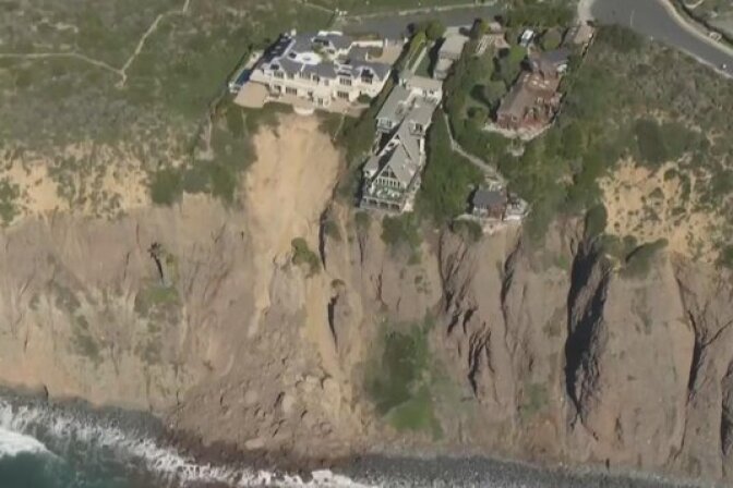 An aerial view shows a large mansion perched on the edge of an eroding seaside cliff