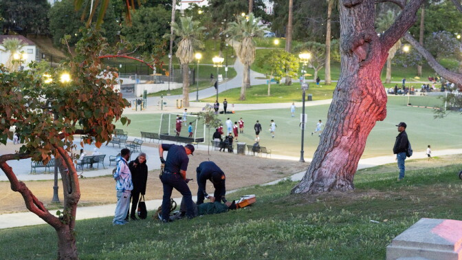 Two paramedics treat a man laying down in the grass. Boys play soccer in the background.