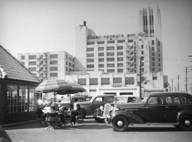 People sit at a table outside, with cars parked nearby and the Sears looming behind them.