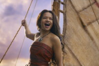 A female-presenting person wearing a terracotta strapless top and a necklace stands holding the rope of a sail. The sail and a cloudy sky are visible in the background.