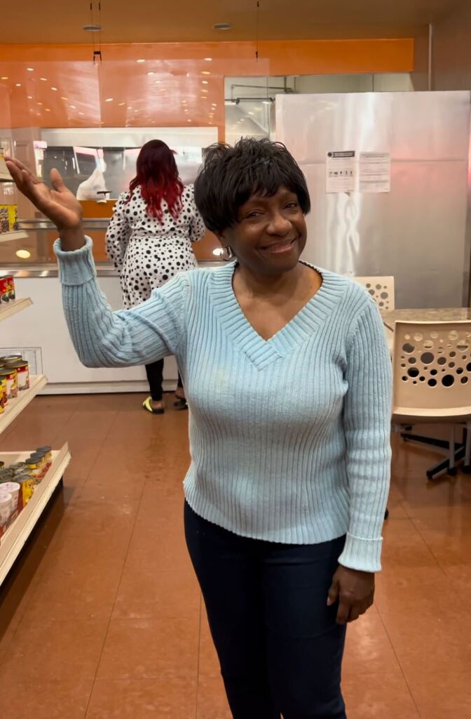 A Black woman with short hair and a light blue long sleeve top and dark pants waves as she stands in a store.