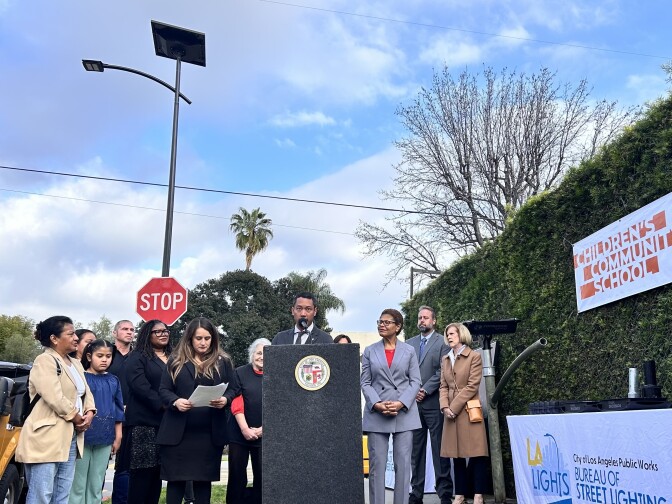 A group of people are gathered around a tall black podium placed on a city sidewalk on a nearly clear, sunny day. A street light, with a solar panel on top, is towering over the group. A man with black hair is speaking into the microphone on the podium. To his right, a table is set up that reads "City of Los Angeles Public Works Bureau of Street Lighting." Above the table, attached to a tall shrub, is an orange and white sign that reads "Children's Community School." 