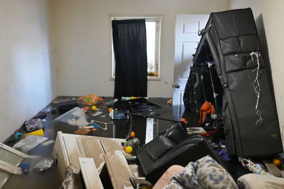 An apartment is flooded and various furniture and household items are floating in a room. Black curtains hang in front of a window in the background.