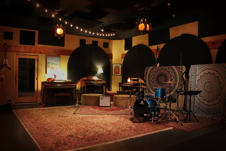The room features soft lighting all around, carpets on the ground and on panels, a drum kit, several keyboards, and other recording equipment. A Flying V guitar hangs on the wall.