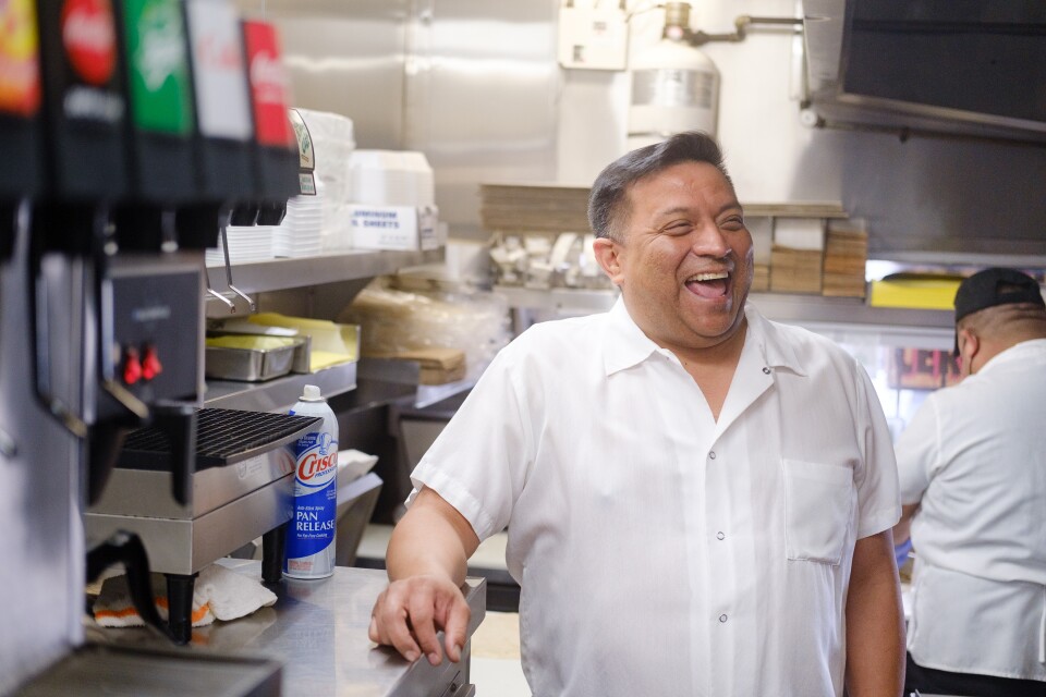 A view of Ryan Carreon standing in a restaurant kitchen from the waist up. He's smiling wide near a drink machine while the crew works behind him.