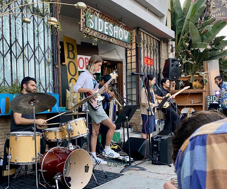 A guy with shoulder-length blond hair plays guitar on sidewalk in front of Sideshow Books surrounded by a drummers and two other people on guitar