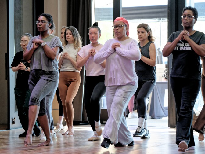 A diverse group of people in a dance studio stand in a staggered formation, practicing a movement together. They are all stepping forward with one foot while holding their hands clasped near their chests, focused and looking ahead.