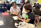 A young person with medium skin tone sits at a black table looking over a piece of paper. Next to them, a young woman with medium skin tone watches, with a sign next to her that says "Roxanna."
