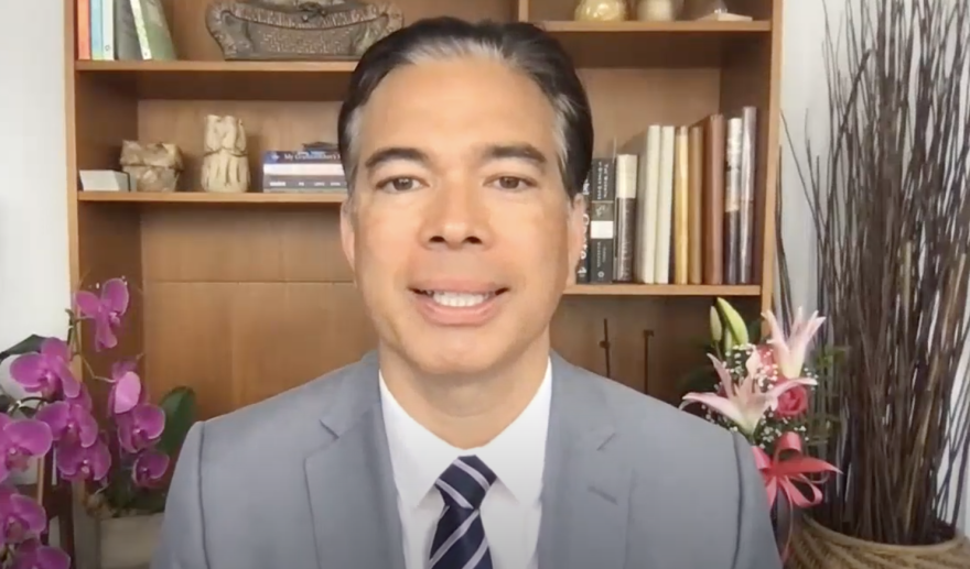 Rob Bonta looks straight at the camera, his mouth slightly open, teeth exposed. He wears a white shirt, blue tie with gray diagonal strips, and grey jacket. Behind him is a purple orchid to the viewer's left and pink and purple flowers to the viewer's right. Also behind him is a cabinet with one shelf filled with books another shelf with books and knicknacks, and another one with knicknacks.