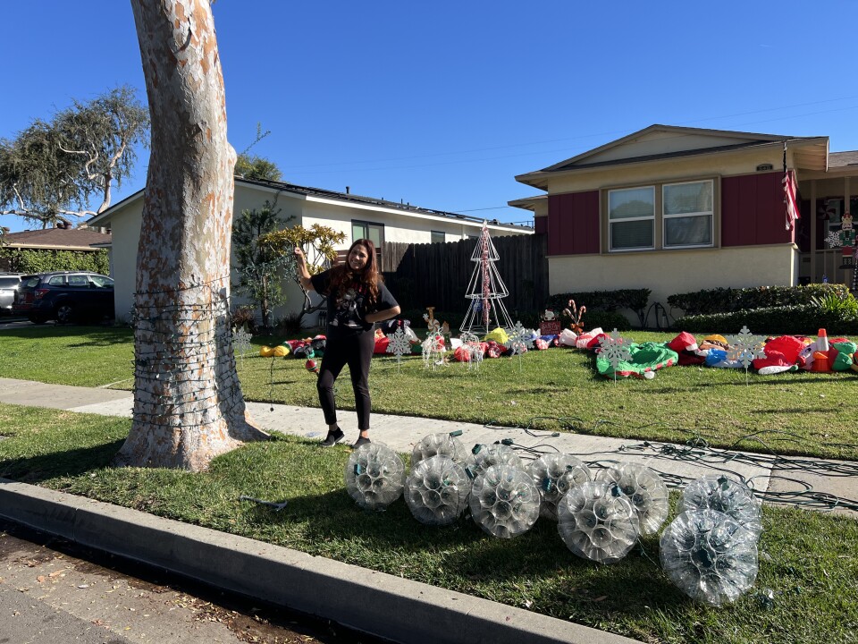 A woman wearing a black tshirt and leggings strings holiday lights around a tree. Other Christmas decorations are visible on her front yard. 