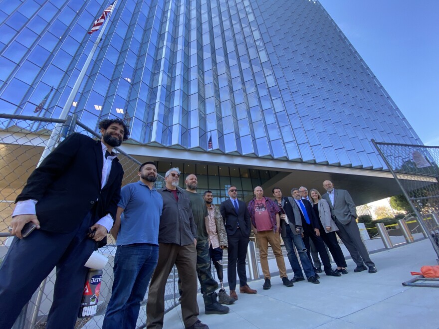 A group of 12 people stand in a semicircle front of a tall glass building with an American flag out front.