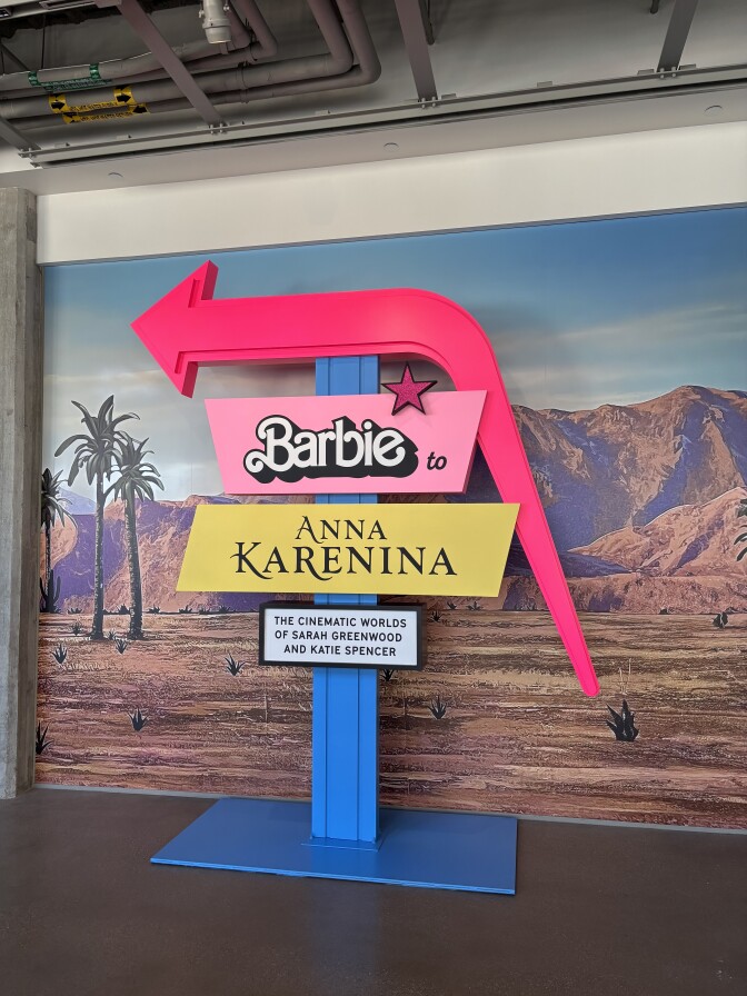 A 3D sign for the "Barbie to Anna Karenina" exhibition at the Academy Museum. At top is a curved hot pink arrow pointing left. It's held up by a bright blue stand. Below the arrow is a pink rectangular sign that reads "Barbie to" in the toy's famous font, and below that a muted yellow sign that says "Anna Karenina" in a more romantic, classic font. At the bottom is a black and white box that reads "The Cinematic Worlds of Sarah Greenwood and Katie Spencer." In the background of all is a desert scene backdrop.