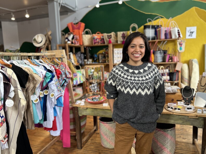 A young woman with light brown skin and dark brown hair wears a grey sweater with white pattern across the front. She smiles in a store surrounded by colorful clothing and goods.
