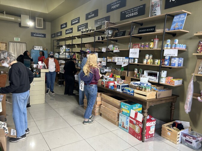 Several people browse tables and shelves stacked high with household and personal goods, including diapers, toilet paper and bedding. 