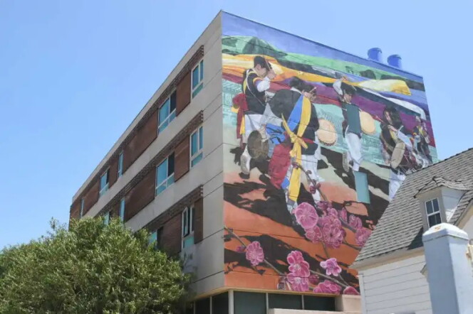 A tall affordable-housing building with a mural covering its upper side wall. The mural depicts Korean drummers in traditional dress mid-performance beneath sweeping ribbons of color, with green mountains and faint architecture in the background and large pink plum blossoms across the lower portion. Green trees and a peaked rooftop are in the foreground under a clear blue sky.
