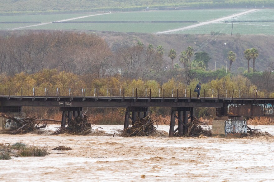 A tumultuous muddy river flows towards a bridge with train tracks. The stands of the bridge in the water are full of drift wood and debris.
