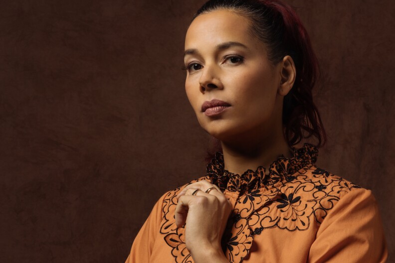 Rhiannon Giddens looks into the camera while her right hand holds the orange sleeve of her blouse. The background is a brown gradient pattern.