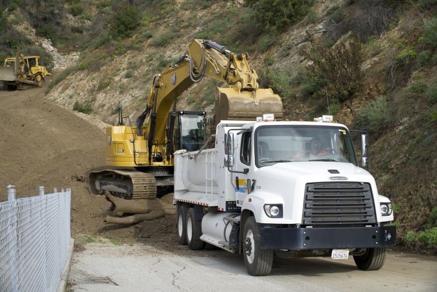 A yellow backhoe pours dirt into the bed of a white dump truck in a debris basin.