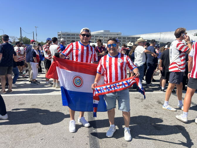 Two men stand outside a stadium and hold flags. One flag reads "PARAGUAY"