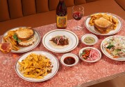 A vibrant, retro-inspired lunch spread set atop a glossy pink cracked-ice Formica table, surrounded by a warm-toned booth featuring  stacked chicken salad sandwich served with golden fries, a single taco , a double-decker patty melt and in the bottom left a plate of loaded chili cheese fries. 