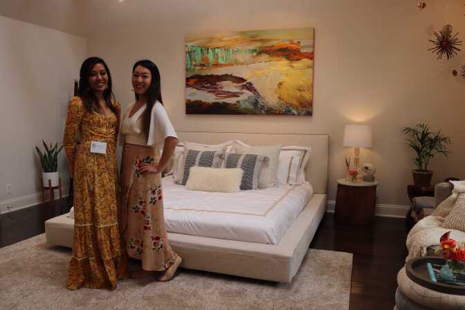 Two women who appear to be in their 20s, both with long black hair, stand smiling and looking at the camera. Behind them is a low bed dressed in white bedsheets, and a colorful painting hung above it. 