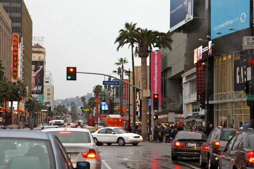 The intersection of Hollywood and Highland is busy with cars and pedestrians. A red street light is seen in the distance. Signs for retail stores like the Gap line the street, as well palm trees.