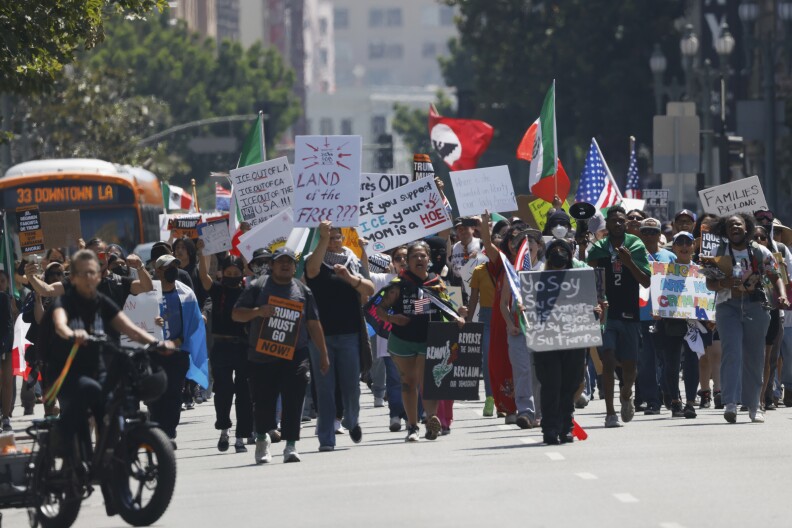 Crowds of people hold flags and signs, with some saying 'Land of the Free?". The people are walking along a street. 