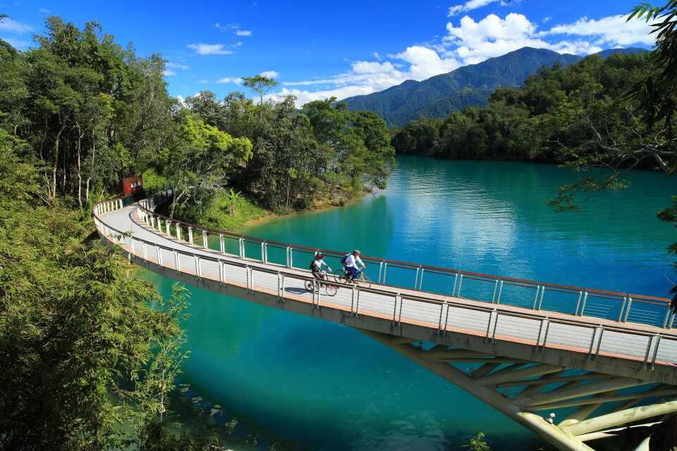 A bicycle path bridge with two bicyclists riding along it, runs over a river in Taiwan. Blue sky with white puffy clouds above. 