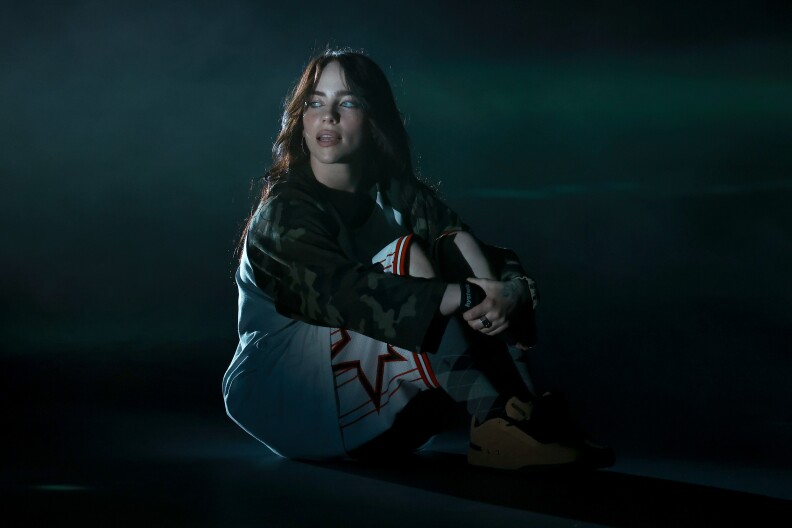 Billie Eilish sits on a stage with his legs folded up, looking away from camera.