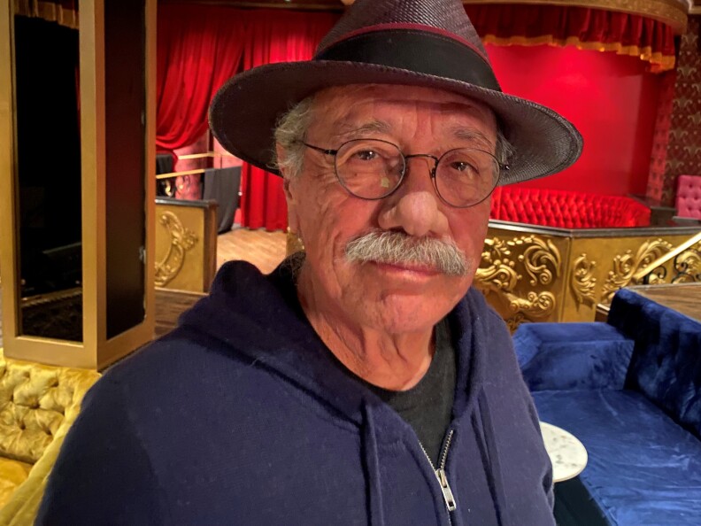 A man wears a brimmed hat and a zip-up hoody in front of a stage in the background with red curtains and gold accents.