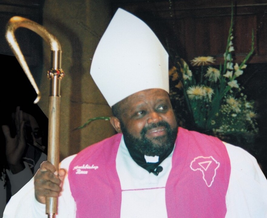 Archbishop Carl Bean is visible from his head to just below his shoulders. He's looking slightly to his left, his hand grasping a golden sceptre with a curved top. He has a white conical hat, white vestments, and pink fabric draped over his shoulders. On the pink fabric, a map of Africa with a triangle in it is visible on his left shoulder.