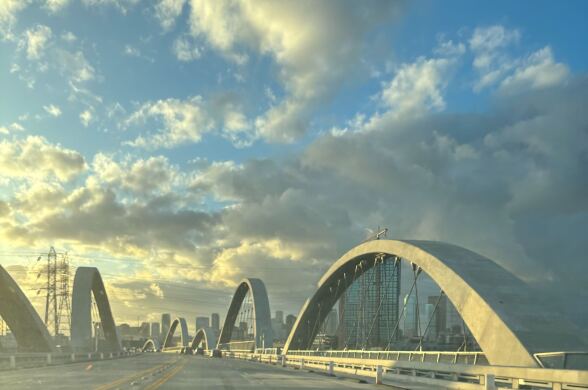 Sunlight reflects off the arches of a bridge with a view of storm clouds and a downtown skyline. 