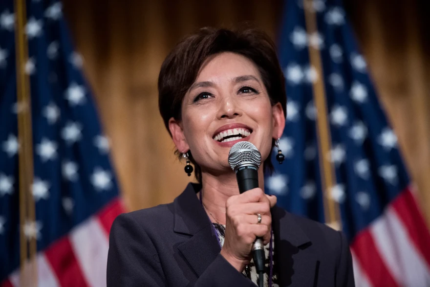 Republican Rep. Young Kim smiles while speaking into a microphone, standing in front of two American flags.