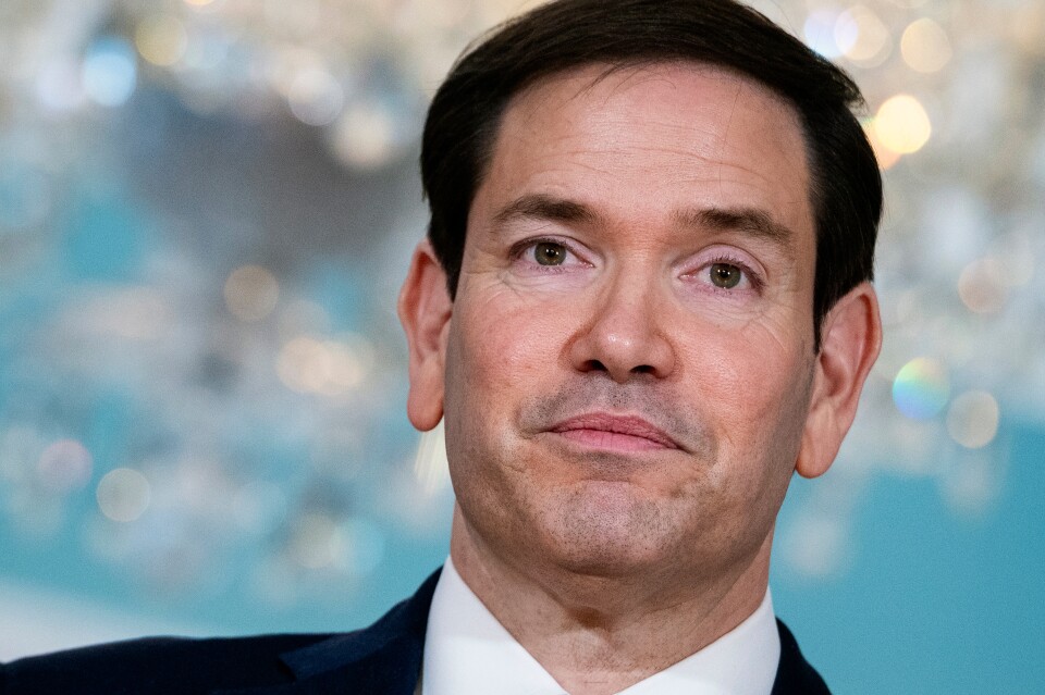 A close up of Secretary of State Marco Rubio, a man with light skin tone, wearing a black suit, white shirt, and tie, looking out with a blue background that is out of focus.